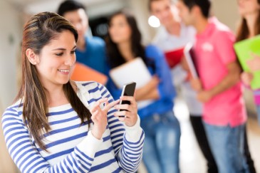 Student looking at her phone in school hallway with students holding notebooks behind her.